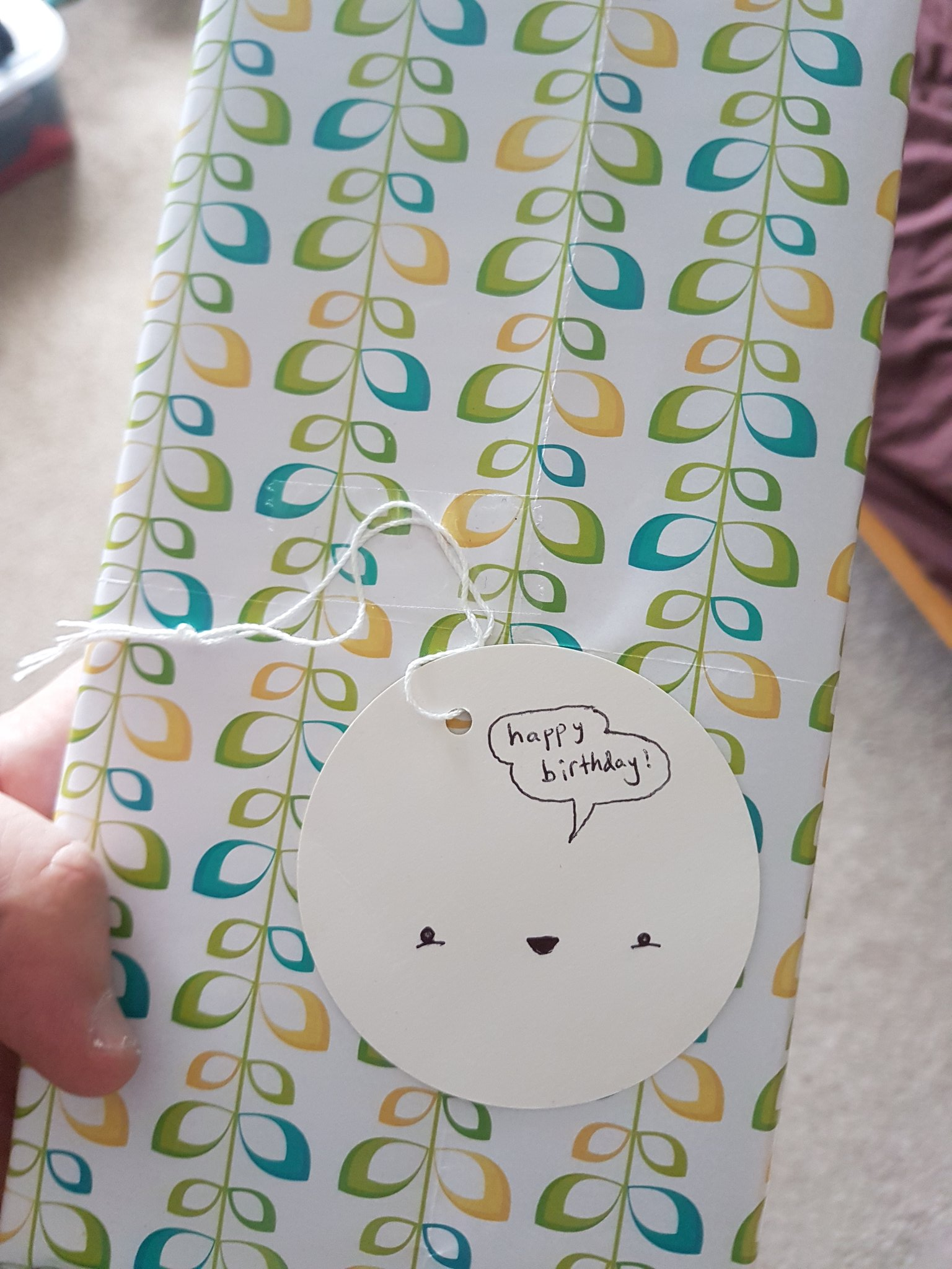 A photograph of a rectangular gift wrapped in paper with a design resembling leaves on plant stems in yellow, green and blue, with a white, circular gift tag in the middle. The tag has a smiling small robot style face drawn on it and a speech bubble that reads "happy birthday!".