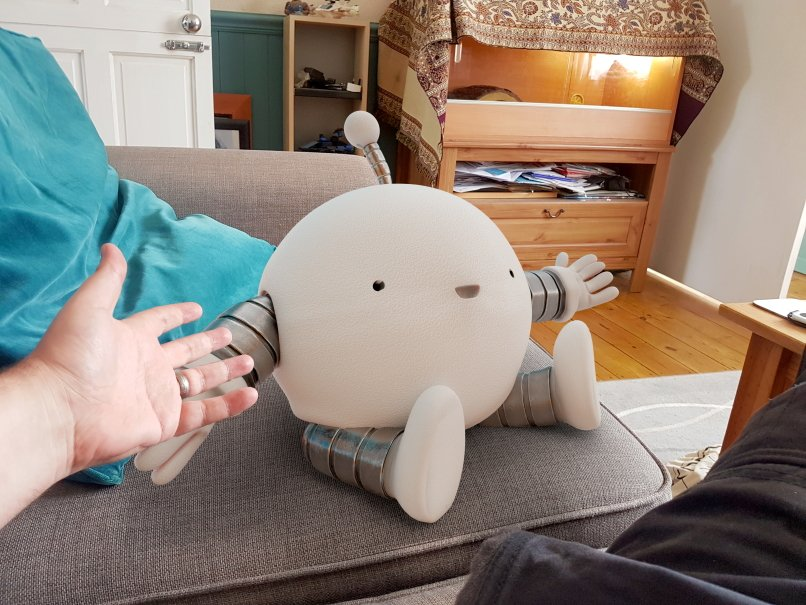 A photograph of a spherical robot with banded arms and legs and an antenna sitting on the sofa in our living room. Our hand is visible in shot, gesturing towards it as the robot holds out its arms and smiles.