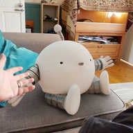 A photograph of a spherical robot with banded arms and legs and an antenna sitting on the sofa in our living room. Our hand is visible in shot, gesturing towards it as the robot holds out its arms and smiles.